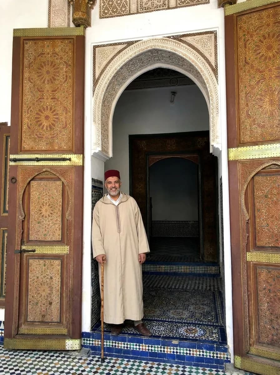 A guide in traditional Moroccan djellaba and fez standing in a carved doorway at Bahia Palace