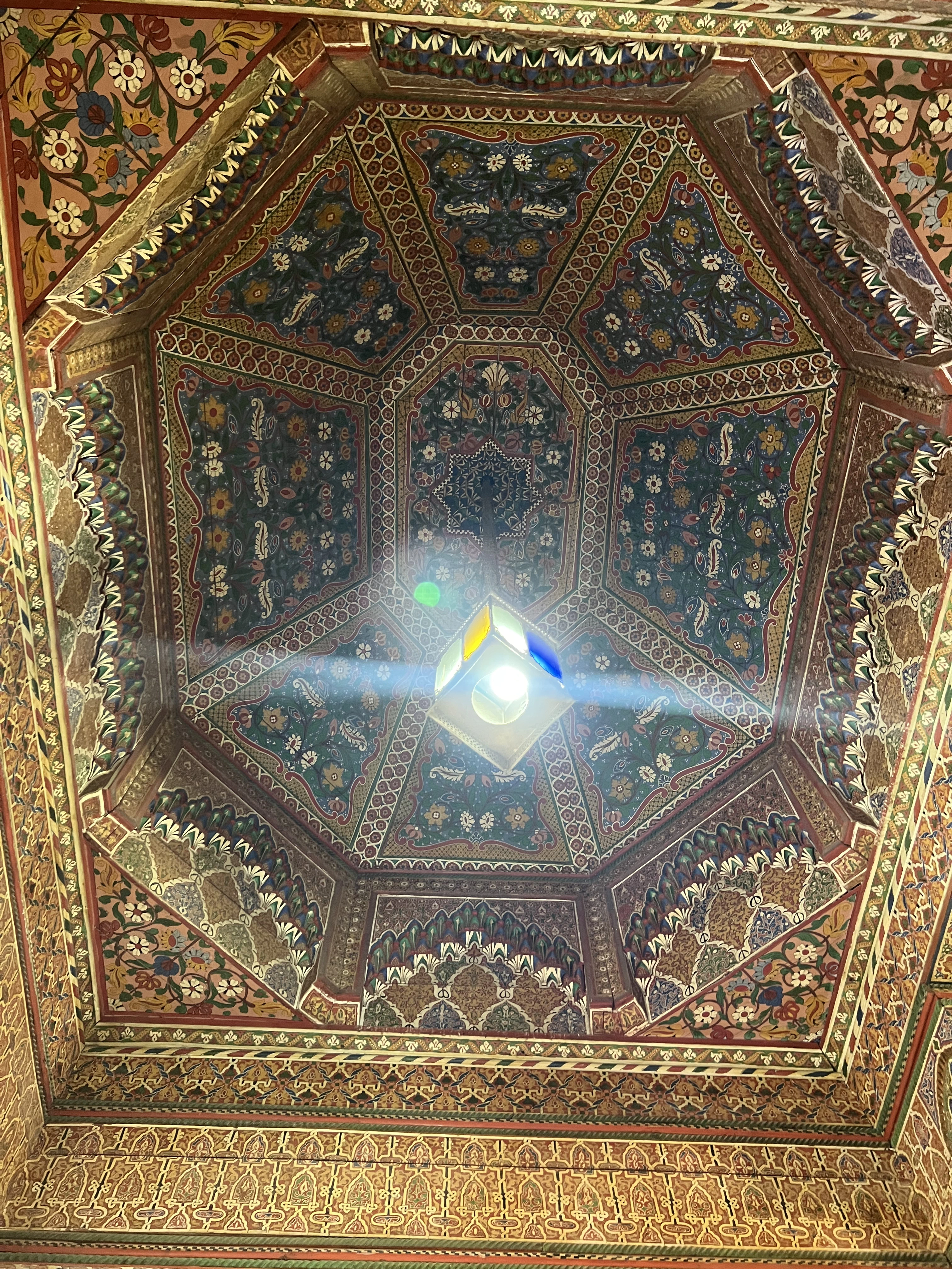 سقف القبة المثمّنة من الأرز المرسوم داخل قصر الباهية بمراكش — نقوش نباتية وهندسية دقيقة بالأحمر والأخضر والذهبي مع كوة مركزية
